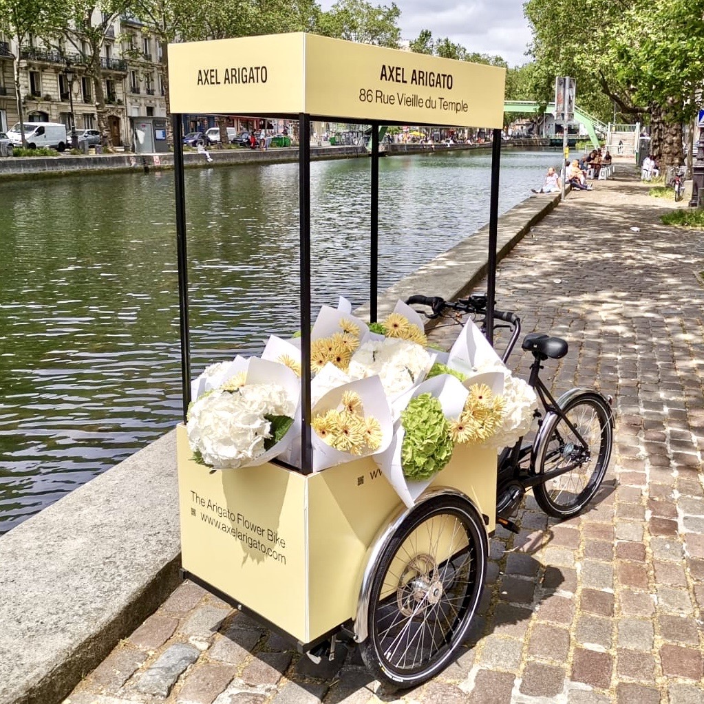Velo Expo pour Axel Arigato Paris Marais. Affichage mobile, animation drive-to-store, distribution de fleurs avec vélo triporteur corgo personnalisé. Campagne communication ooh hors-media par activation affichage mobile digital avec vélo triporteur publicitaire street marketing | Velo Expo | Agence Veymont